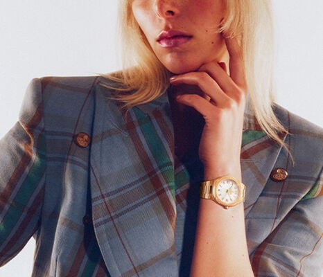 Close-up of model wearing Vivienne Westwood gold bracelet watch with white dial, styled with tartan blazer.